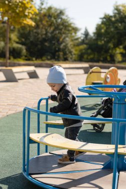 Gri şapkalı ve siyah deri ceketli küçük mutlu çocuk sonbaharda dışarıda oyun alanında oynuyor.