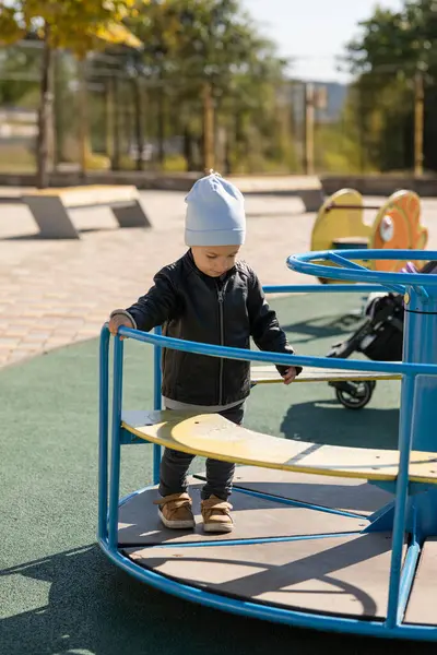 Gri şapkalı ve siyah deri ceketli küçük mutlu çocuk sonbaharda dışarıda oyun alanında oynuyor.
