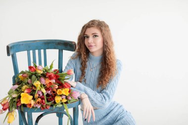 Kırmızı kıvırcık saçlı güzel bir genç kız ve beyaz bir arka planda, yumuşak mavi bir elbiseyle kocaman bir buket çok renkli laleyle bir fotoğraf stüdyosunda kocaman gözleri var. Fotoğraf mankeni. Model.