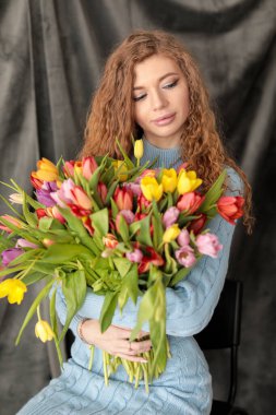 Kırmızı kıvırcık saçlı, güzel, genç, ince bir kız ve gri arka planda, yumuşak mavi bir elbise içinde, bir fotoğraf stüdyosunda bir buket çok renkli lalesi olan, kocaman gözleri olan bir kız. Fotoğraf mankeni. Model.