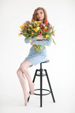 Kırmızı kıvırcık saçlı güzel bir genç kız ve beyaz bir arka planda, yumuşak mavi bir elbiseyle kocaman bir buket çok renkli laleyle bir fotoğraf stüdyosunda kocaman gözleri var. Fotoğraf mankeni. Model.