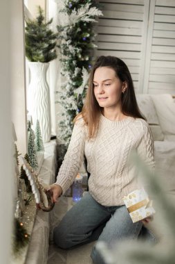 Pencere kenarında beyaz bir fotoğraf bölgesinde beyaz kazak ve kot pantolon giyen genç, güzel bir kız. Fotoğraf mankeni. Modellik. Yeni yıl. Noel zamanı. Kış.