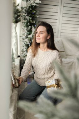 Pencere kenarında beyaz bir fotoğraf bölgesinde beyaz kazak ve kot pantolon giyen genç, güzel bir kız. Fotoğraf mankeni. Modellik. Yeni yıl. Noel zamanı. Kış.