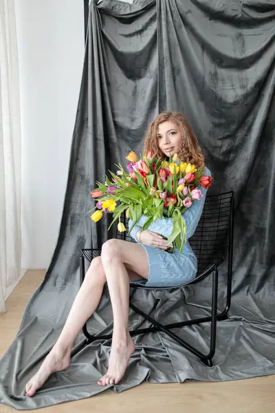 Kırmızı kıvırcık saçlı, güzel, genç, ince bir kız ve gri arka planda, yumuşak mavi bir elbise içinde, bir fotoğraf stüdyosunda bir buket çok renkli lalesi olan, kocaman gözleri olan bir kız. Fotoğraf mankeni. Model.