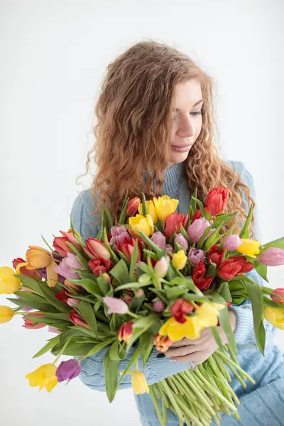 Kırmızı kıvırcık saçlı güzel bir genç kız ve beyaz bir arka planda, yumuşak mavi bir elbiseyle kocaman bir buket çok renkli laleyle bir fotoğraf stüdyosunda kocaman gözleri var. Fotoğraf mankeni. Model.