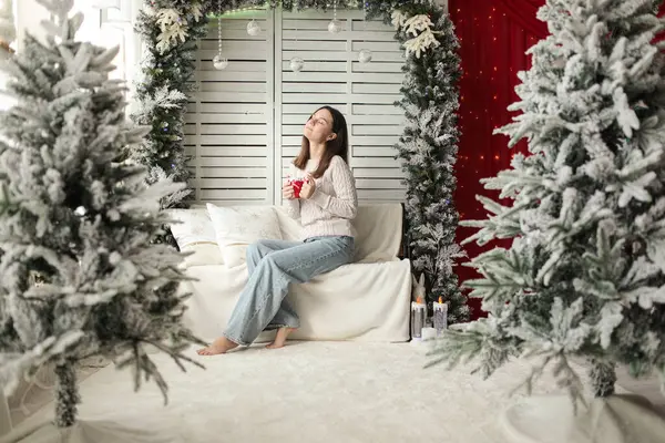 Pencere kenarında beyaz bir fotoğraf bölgesinde beyaz kazak ve kot pantolon giyen genç, güzel bir kız. Fotoğraf mankeni. Modellik. Yeni yıl. Noel zamanı. Kış.