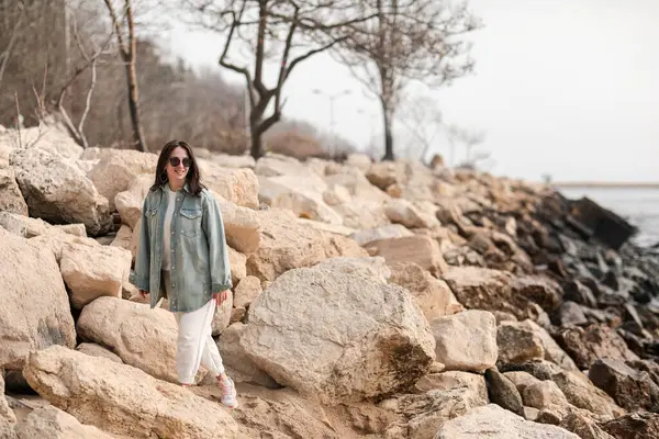 Koyu renk saçlı, kot ceketli ve beyaz pantolonlu güzel bir kız sahilde yürüyor..