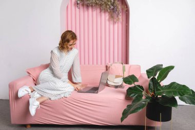 Pembe elbiseli bir kadın beyaz bir odada bir buket lale ile yatakta oturuyor..