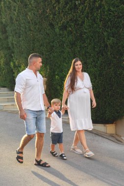 Mutlu genç bir aile, beyaz giysiler içinde, yazın parkta yürüyor. Annelik. Anne ve oğlu. Baba ve oğul. Anne, baba ve oğul. - Aile. Anneler Günü. Babalar Günü. Hamilelik. Hamile kız.