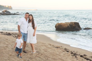 Mutlu genç bir aile, beyaz giysiler içinde, yazın sahilde. Annelik. Anne ve oğlu. Baba ve oğul. Anne, baba ve oğul. - Aile. Anneler Günü. Babalar Günü. Hamilelik. Hamile kız.