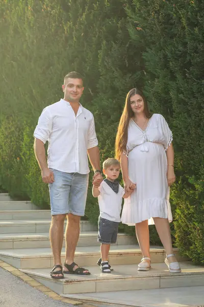 Mutlu genç bir aile, beyaz giysiler içinde, yazın parkta yürüyor. Annelik. Anne ve oğlu. Baba ve oğul. Anne, baba ve oğul. - Aile. Anneler Günü. Babalar Günü. Hamilelik. Hamile kız.