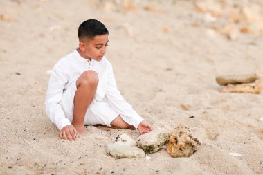 Beyaz elbiseli çocuk kumsalda oynuyor. Denizde, kumların üzerinde. Yazın deniz kabukları ve taşlarla.