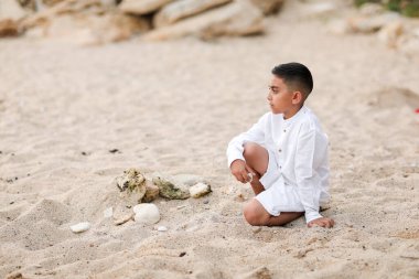 Beyaz elbiseli çocuk kumsalda oynuyor. Denizde, kumların üzerinde. Yazın deniz kabukları ve taşlarla.