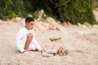 Beyaz elbiseli çocuk kumsalda oynuyor. Denizde, kumların üzerinde. Yazın deniz kabukları ve taşlarla.