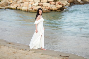 Yazın kumsalda, uzun siyah saçlı, beyaz, uzun elbiseli güzel bir kız.