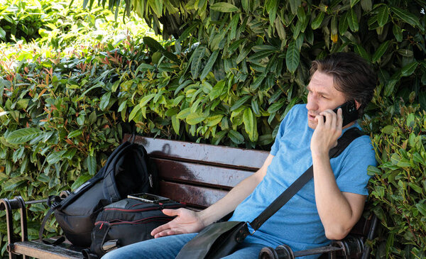 серьезный молодой человек, сидящий на скамейке и разговаривающий по телефону в парке