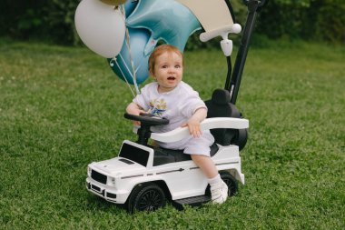 Küçük bebek, balonları çimlerin üzerine park etmiş beyaz bir arabada oturuyor. Ufaklık. Yaz.