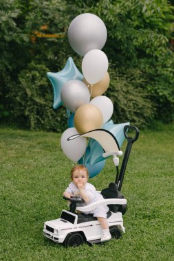 Küçük bebek, balonları çimlerin üzerine park etmiş beyaz bir arabada oturuyor. Ufaklık. Yaz.