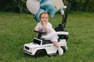 Küçük bebek, balonları çimlerin üzerine park etmiş beyaz bir arabada oturuyor. Ufaklık. Yaz.