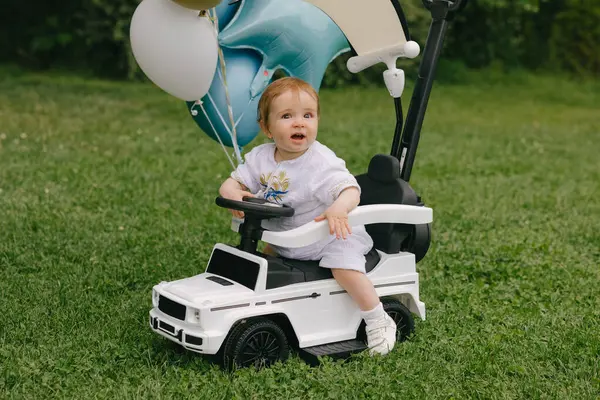 Küçük bebek, balonları çimlerin üzerine park etmiş beyaz bir arabada oturuyor. Ufaklık. Yaz.