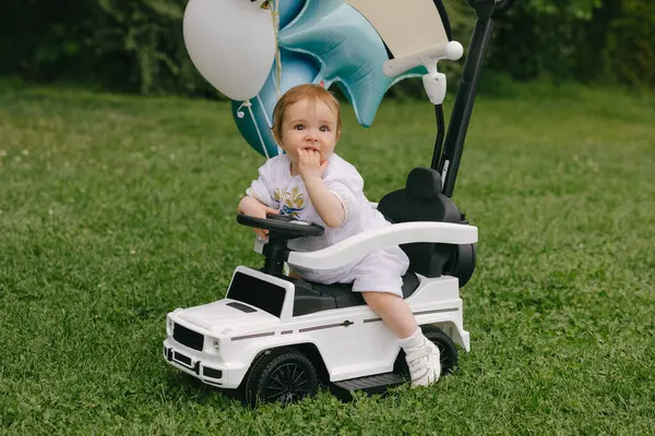 Küçük bebek, balonları çimlerin üzerine park etmiş beyaz bir arabada oturuyor. Ufaklık. Yaz.