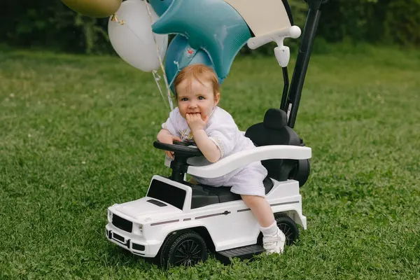 Küçük bebek, balonları çimlerin üzerine park etmiş beyaz bir arabada oturuyor. Ufaklık. Yaz.