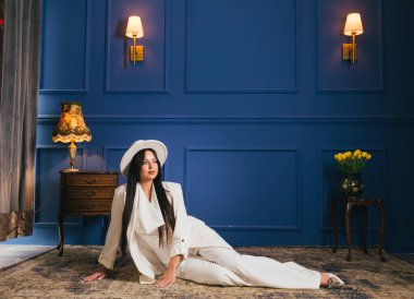 Uzun siyah saçlı, beyaz takım elbiseli ve mavi arka planda şapkası olan güzel bir genç kız. Esmer. İş kadını.