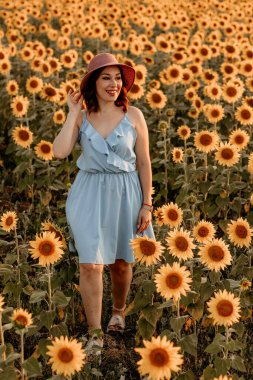 Güzel, mutlu, mavi elbiseli ve şapkalı genç kız gün batımında ayçiçeği tarlasında
