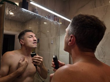 Adamın biri banyoda sakalını ve bıyığını aynanın önünde tıraş ediyor.