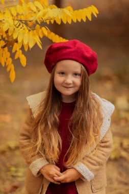 Kırmızı bereli ve sonbahar parkında açık kahverengi ceketli mutlu küçük kız. Sonbahar konsepti. Sonbahar havası sonbahar