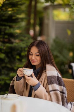 Güzel, mutlu, esmer bir kız sonbahar parkında battaniyeye sarılı bir kafede oturur ve bir fincan kahve içer. Sonbahar konsepti. Sonbahar sezonu. Sonbahar. konfor
