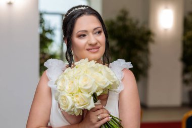 A lovely bride smiling while holding a bouquet of cream roses, standing indoors in a ceremony room with flags visible in the background