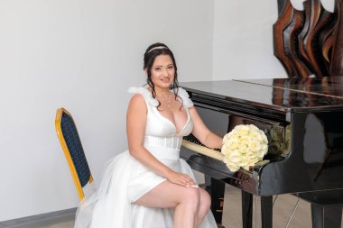 A beautiful bride in a short wedding dress with a tulle skirt, sitting next to a black grand piano, holding a bouquet of cream roses