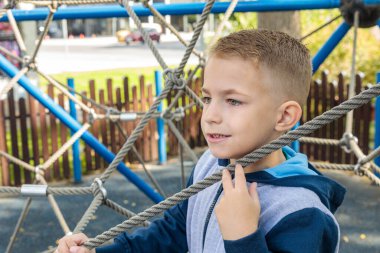 Cute boy with a modern haircut playing and climbing on a rope net structure at an outdoor playground, looking away, close-up