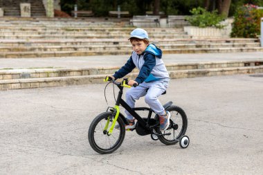 A young boy in a cap and hoodie is riding a black and neon green children's bicycle with training wheels on a paved square with stone bleachers in the background