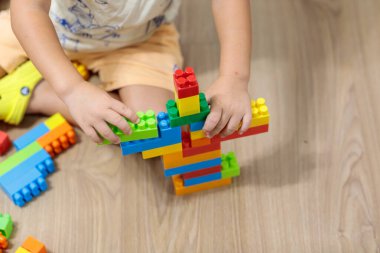 Unrecognizable child sitting on a wooden floor, actively engaged in building a tall, colorful structure with plastic construction blocks, showcasing creative play and early learning
