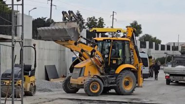 Yellow JCB Backhoe Loader on a Construction Site Filling a Dump Truck with Gravel Near a Concrete Wall and Other Heavy Equipment