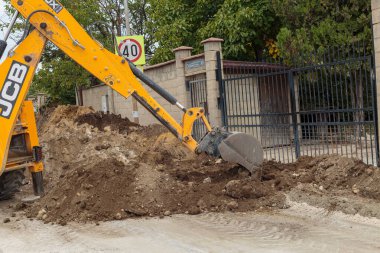 Yellow JCB backhoe loader bucket digs up a pile of brown dirt and earth on an unpaved road next to a fence during groundwork and construction. 
