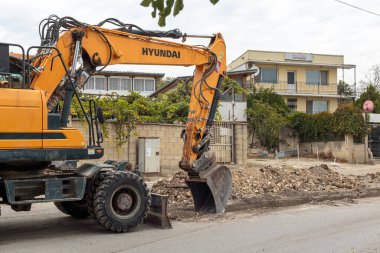 Yellow JCB backhoe loader bucket digs up a pile of brown dirt and earth on an unpaved road next to a fence during groundwork and construction. 