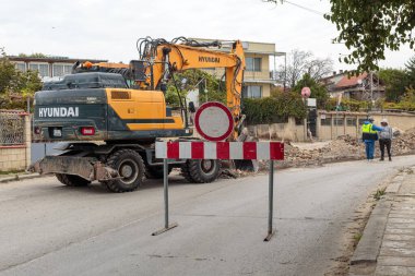 Yellow wheeled Hyundai excavator digging a trench on an urban road next to two construction workers, illustrating municipal groundwork and repair.  Varna, Bulgaria. - 1.10.2025