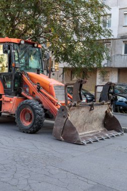 Bright orange backhoe loader parked on a suburban street in front of a residential apartment building, ready for construction or road repair work.