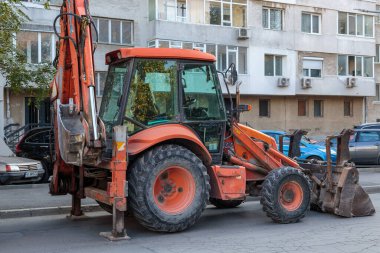 Bright orange backhoe loader parked on a suburban street in front of a residential apartment building, ready for construction or road repair work.