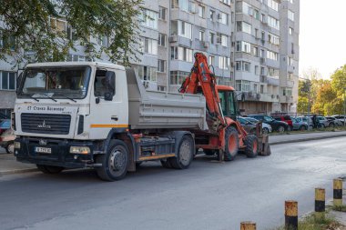 Heavy dump truck carrying a backhoe loader on a suburban residential street in front of a large apartment building during city construction or infrastructure maintenance.
