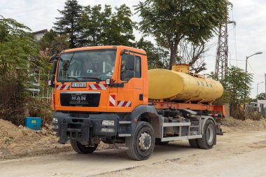 Orange MAN tanker truck with a yellow water tank drives on an unpaved road at a construction site in a residential area