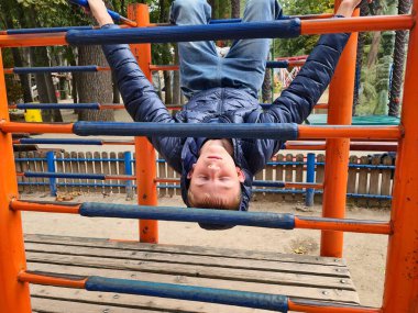 teenager in a blue puffer jacket and jeans posing on a colorful metal climbing frame at a park playground on a cloudy day, enjoying outdoor activity and childhood