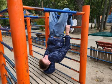 A boy dressed in a blue puffer jacket and jeans hangs upside down from the orange metal bars of a playground climbing frame, having fun in the park