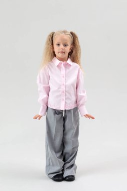 Full-length studio portrait of a young girl with blonde pigtails, standing and posing seriously in a pink shirt and wide grey pants.  Model, photo model, posing, photo studio