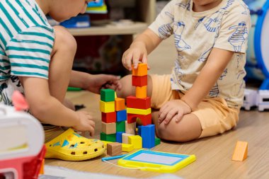Two little boys playing on the floor with colorful construction blocks in a messy living room, surrounded by toys and a TV stand