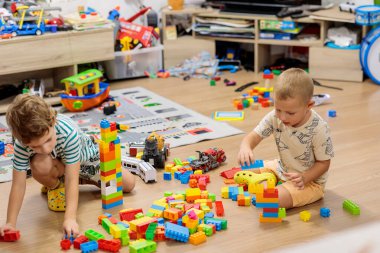 Two little boys playing on the floor with colorful construction blocks in a messy living room, surrounded by toys and a TV stand