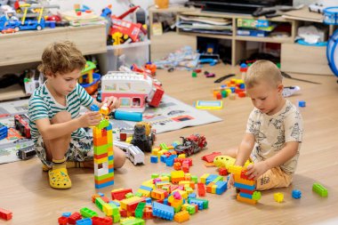 Two little boys playing on the floor with colorful construction blocks in a messy living room, surrounded by toys and a TV stand